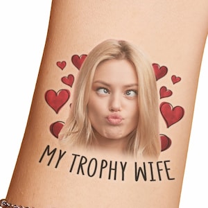 Pode incluir: Uma tatuagem temporária com o rosto de uma mulher e o texto "MY TROPHY WIFE" rodeado de corações vermelhos.