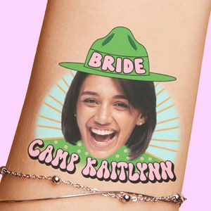 Puede incluir: Un tatuaje temporal que representa a una mujer con un sombrero verde de guardabosques con la palabra "BRIDE" en él. El tatuaje también incluye el texto "CAMP KAITLYNN" en una fuente retro.