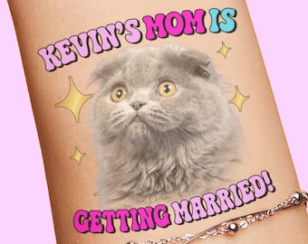 Puede incluir: Un tatuaje temporal que muestra un gato Scottish Fold gris con ojos amarillos, rodeado de iconos de estrellas doradas. El texto dice KEVIN'S MOM IS en rosa y azul, y GETTING MARRIED! en rosa, con una fina pulsera de cadena plateada debajo.