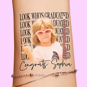 Può includere: Un tatuaggio temporaneo con un bambino con occhiali da sole rosa e una chitarra. Il tatuaggio include il testo "LOOK WHO'S GRADUATED" e "Congrats Sophia". Sotto è visibile un braccialetto d'argento.