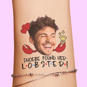 May include: A temporary tattoo featuring a smiling person's face surrounded by two red hearts, red lobster claws holding a yellow diamond ring, and the text PHOEBE FOUND HER LOBSTER with colourful dots between the letters of the word LOBSTER.