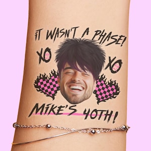 May include: A temporary tattoo on an arm with the text "IT WASN'T A PHASE!" and "MIKE'S 40TH!" featuring a smiling man's face, pink and black checkered hearts, and "XO" symbols. A silver bracelet is visible.