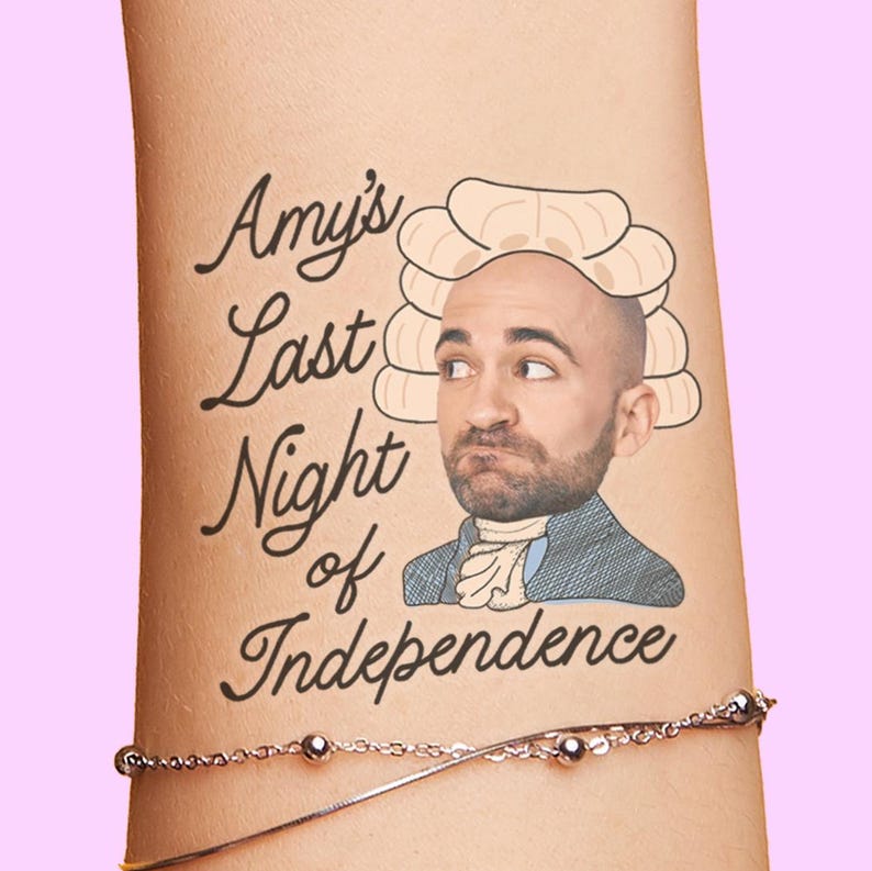 May include: A humorous tattoo on an arm features the text "Amy's Last Night of Independence" in elegant script. The design includes a cartoon illustration of a man in a powdered wig and colonial attire, set against a pink background.