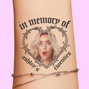 May include: A temporary tattoo featuring a woman's portrait within a heart-shaped barbed wire design. The text "in memory of ashley's twenties" is arched around the heart. A silver bracelet with small beads is visible below the tattoo.