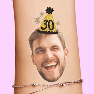 Op de afbeelding: Een tijdelijke tatoeage met een realistisch fotografisch portret van een lachend persoon met een baard, die een gouden glitterfeestmuts draagt met het getal 30 in het zwart gedrukt, geaccentueerd door kleine zwarte sterillustraties.