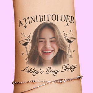 May include: Temporary tattoo with the text "A Tini Bit Older" above a smiling woman's face, with two martini glasses on either side. Below the face is the text "Ashley's Dirty Thirty". A silver bracelet is visible below the tattoo.