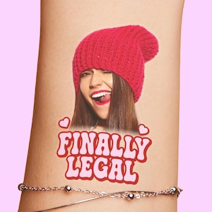 May include: A temporary tattoo featuring a smiling woman wearing a red knit beanie. The text "FINALLY LEGAL" is displayed in a pink, bubble-style font, with two small hearts above the text. A silver bracelet is visible.
