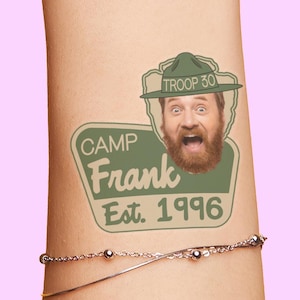 May include: A temporary tattoo featuring a man's face with a beard and a green hat that says "Troop 30." Below the face is a green sign that says "Camp Frank Est. 1996."