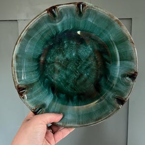 May include: A teal-coloured ashtray with a glossy finish. The ashtray has four notched rests for cigarettes and a dark brown interior. The rim is a lighter shade of teal. The ashtray is held by a hand.