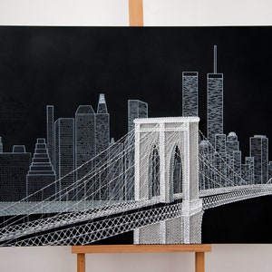May include: A black and white string art depiction of the Brooklyn Bridge and the New York City skyline. The bridge is in the foreground and the skyline is in the background. The art is created with white string on a black canvas.