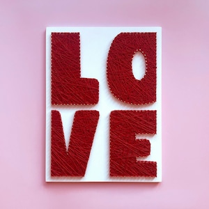 以下が含まれることがあります： 白い長方形の看板に、赤い糸で「LOVE」の文字が描かれています。文字は赤い糸で埋められ、白い背景に配置され、ピンク色の背景になっています。