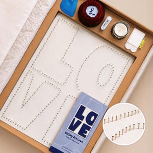 Pode incluir: Um kit de arte com fios numa caixa de cartão contendo um quadro branco com pregos dispostos para soletrar LOVE, um novelo de fio vermelho escuro com etiqueta L, uma ferramenta azul, um cortador branco, uma vela e um manual String Art Love.