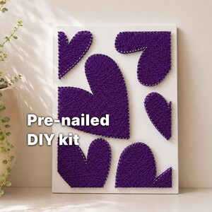Op de afbeelding: Een wit houten bord met een string art ontwerp van paarse harten. De harten zijn gemaakt met paars garen gewikkeld rond voorgehamerde contouren. De tekst op het bord luidt "Pre-genageld DIY kit".
