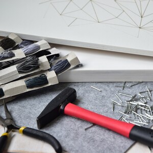 May include: A set of craft supplies for string art, including a white canvas, gray yarn, a hammer, nails, and pliers.