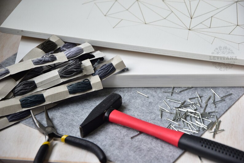 May include: A set of craft supplies for string art, including a white canvas, gray yarn, a hammer, nails, and pliers.