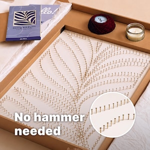 Könnte beinhalten: Ein String-Art-Set in einem Karton mit einer weißen Platte und goldenen Nägeln in Palmenblatt-Optik. Enthält ein dunkelrotes Garnknäuel, eine kleine Kerze im Glas und eine blaue Anleitung mit der Aufschrift String Art Palm.