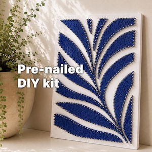 Könnte beinhalten: Ein rechteckiges weißes Brett mit einem vorgenagelten String-Art-Design, bei dem tiefblauer Faden abstrakte blattartige Formen bildet. Der Text Pre-nailed DIY kit ist über das Bild gelegt. Das Brett steht aufrecht auf einer ebenen Fläche.