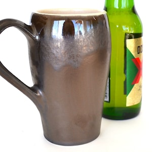 May include: A tall, brown ceramic mug with a large handle, next to a green beer bottle. The mug has a metallic glaze and a cream-colored interior. The beer bottle has a gold label with red and white text.