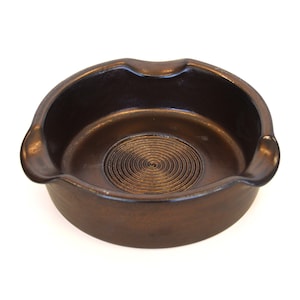 May include: A bronze-colored ashtray with a circular design in the center. The ashtray has two curved rests and a smooth, rounded shape. The interior features a spiral pattern.