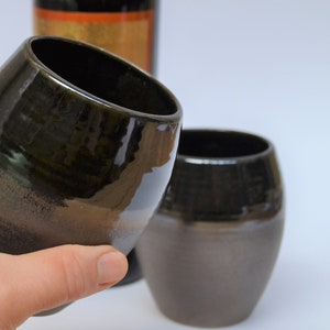 May include: Two black and brown ceramic cups, one being held in a hand. The cups have a glossy finish and a rounded shape.
