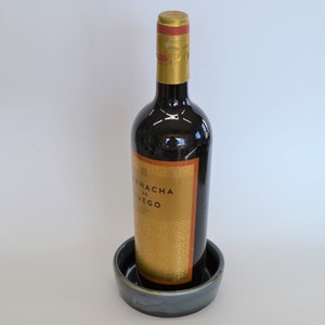 K&ouml;nnte beinhalten: Eine dunkle Glasflasche Wein mit einem goldenen Etikett, auf dem "Fincha de Fuego" steht, steht auf einem blauen und wei&szlig;en Keramikuntersetzer.