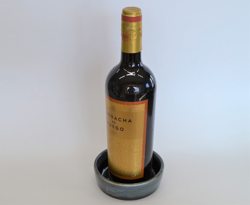 K&ouml;nnte beinhalten: Eine dunkle Glasflasche Wein mit einem goldenen Etikett, auf dem "Fincha de Fuego" steht, steht auf einem blauen und wei&szlig;en Keramikuntersetzer.