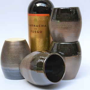 May include: Four dark brown ceramic wine glasses with a metallic sheen, stacked on top of each other. A bottle of wine with a gold label that reads "Garnacha de Fuego" is in the background.