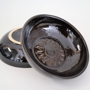 May include: Two black ceramic bowls, one with a sun design, are stacked on a white surface.