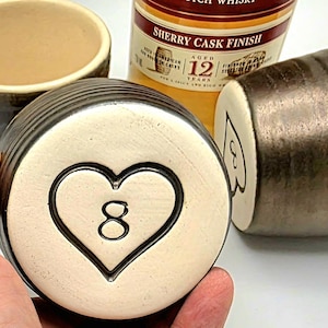 May include: A set of ceramic drinking vessels, including a tumbler and two cylindrical cups. One cup features a heart with the number 8, another has a spade symbol with the number 3. A bottle of Scotch whisky is in the background.