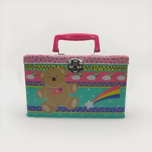 May include: A rectangular metal lunchbox with a pink handle and clasp. The box features a cartoon teddy bear, rainbows, clouds, and a shooting star design. The colour scheme includes pink, teal, yellow, and blue.