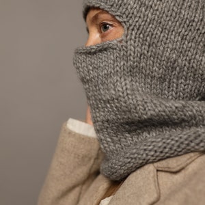 Puede incluir: Una balaclava de punto gris con un dise&ntilde;o de pliegue. La balaclava la lleva puesta una persona con un abrigo marr&oacute;n.