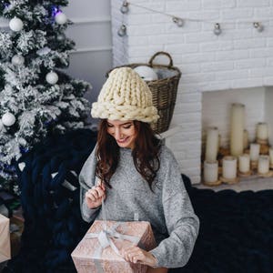 May include: A woman wearing a chunky knit cream hat sits on a blue knitted blanket and opens a gift box. The gift box is wrapped in pink paper with a white ribbon. A Christmas tree is in the background.