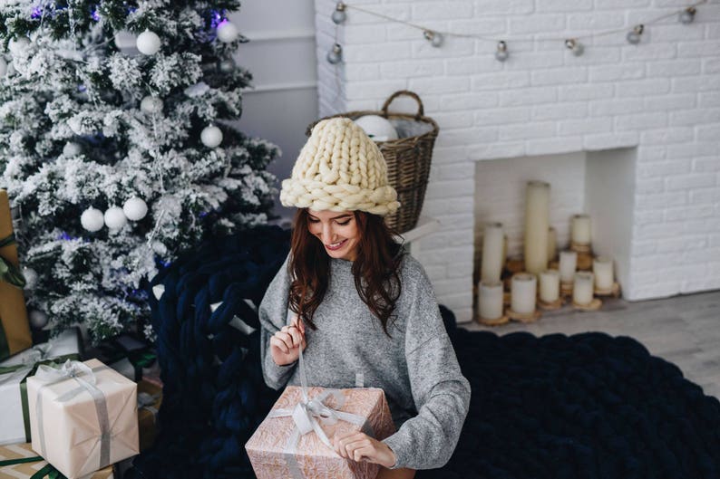 May include: A woman wearing a chunky knit cream hat sits on a blue knitted blanket and opens a gift box. The gift box is wrapped in pink paper with a white ribbon. A Christmas tree is in the background.