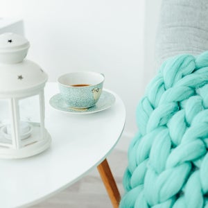 May include: A white lantern with a star design sits on a white table next to a cup of tea and a beige candle. A turquoise chunky knit blanket is draped over a chair in the background.