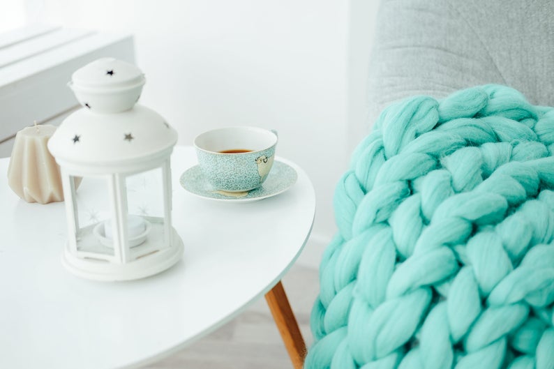 May include: A white lantern with a star design sits on a white table next to a cup of tea and a beige candle. A turquoise chunky knit blanket is draped over a chair in the background.