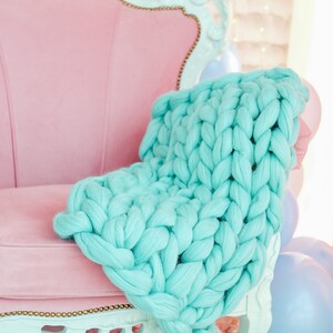 May include: A light blue chunky knit throw draped over a pink velvet armchair with ornate details. The chair is in a room with a white background and blue balloons.