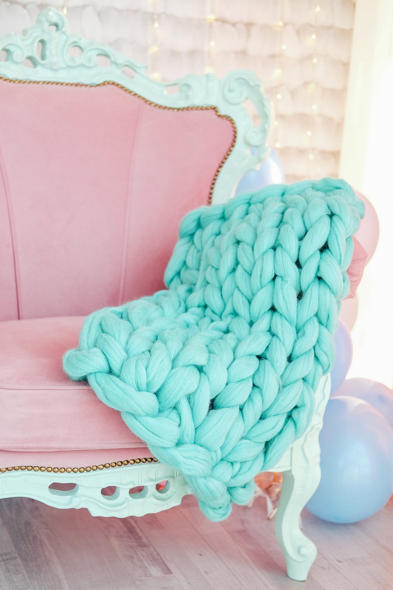 May include: A light blue chunky knit throw draped over a pink velvet armchair with ornate details. The chair is in a room with a white background and blue balloons.