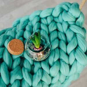 May include: A teal blue chunky knit blanket with a small glass jar containing a green plant and a cork lid.