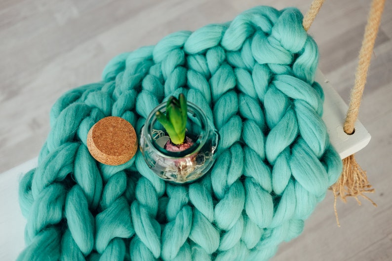 May include: A teal blue chunky knit blanket with a small glass jar containing a green plant and a cork lid.
