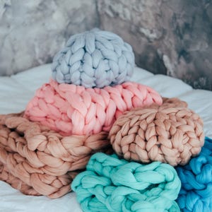 May include: A stack of chunky knit throw pillows in various colors, including light blue, pink, beige, and teal. The pillows are arranged on a white bedspread.