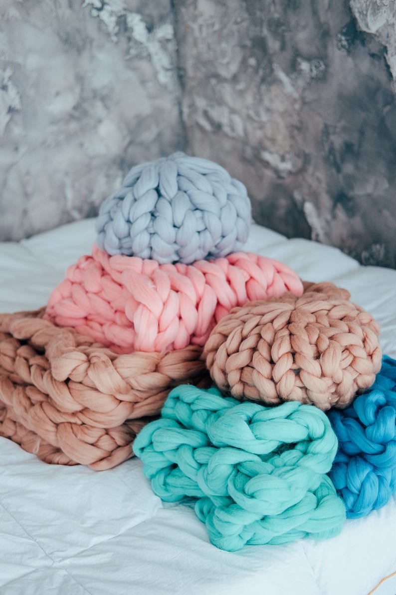 May include: A stack of chunky knit throw pillows in various colors, including light blue, pink, beige, and teal. The pillows are arranged on a white bedspread.