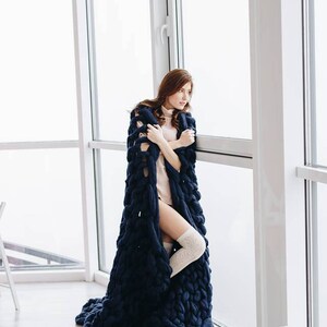 May include: A woman wearing a long, navy blue knitted blanket, standing by a window. She is wearing a light pink turtleneck sweater and white knee-high socks.