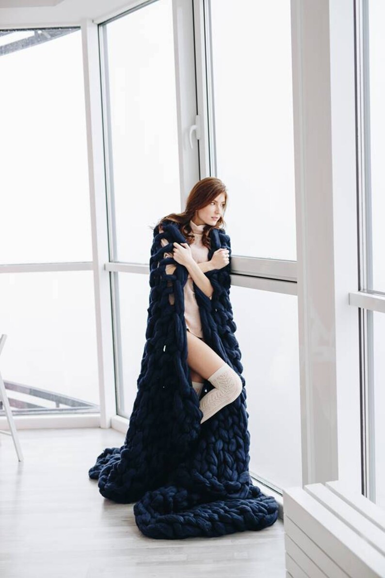 May include: A woman wearing a long, navy blue knitted blanket, standing by a window. She is wearing a light pink turtleneck sweater and white knee-high socks.
