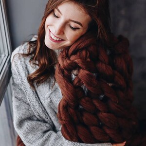 May include: A woman wearing a gray sweater is sitting by a window and holding a large, brown chunky knit blanket.