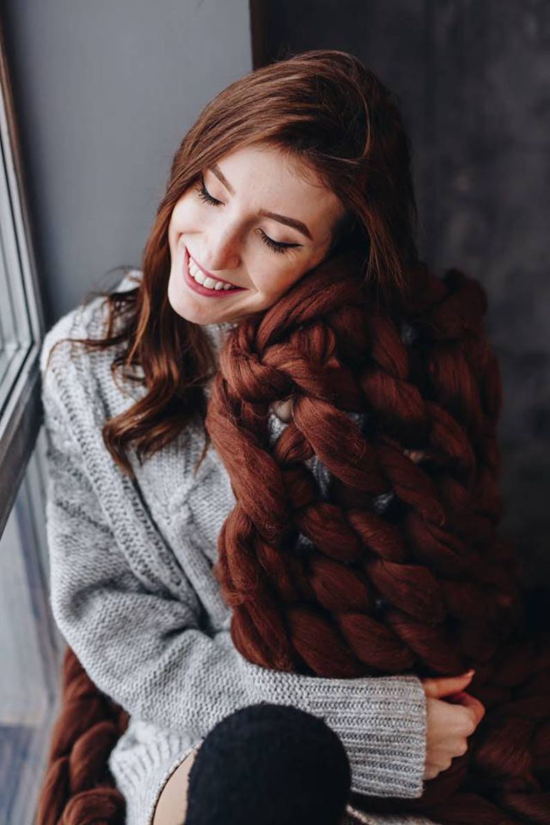 May include: A woman wearing a gray sweater is sitting by a window and holding a large, brown chunky knit blanket.