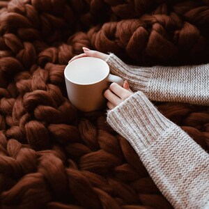 May include: A white ceramic mug held by hands wearing a light grey knitted sweater, resting on a brown chunky knit blanket.