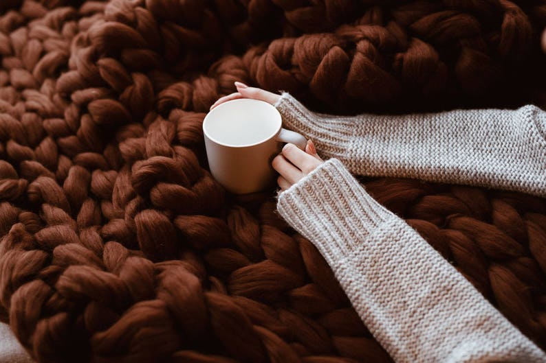 May include: A white ceramic mug held by hands wearing a light grey knitted sweater, resting on a brown chunky knit blanket.