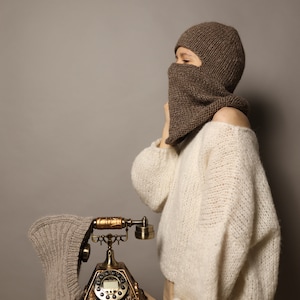 Puede incluir: Una persona con una balaclava de punto marr&oacute;n y un su&eacute;ter de punto blanco sostiene un tel&eacute;fono rotatorio vintage. El tel&eacute;fono est&aacute; sobre una mesa de madera.