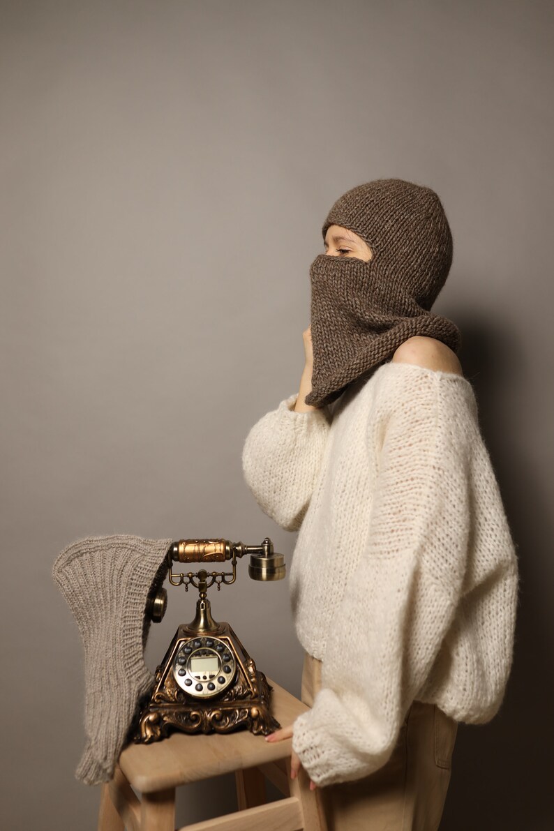 Puede incluir: Una persona con una balaclava de punto marr&oacute;n y un su&eacute;ter de punto blanco sostiene un tel&eacute;fono rotatorio vintage. El tel&eacute;fono est&aacute; sobre una mesa de madera.