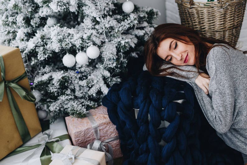 May include: A woman is sleeping on a blue knitted blanket with a Christmas tree in the background. There are wrapped gifts in the foreground.
